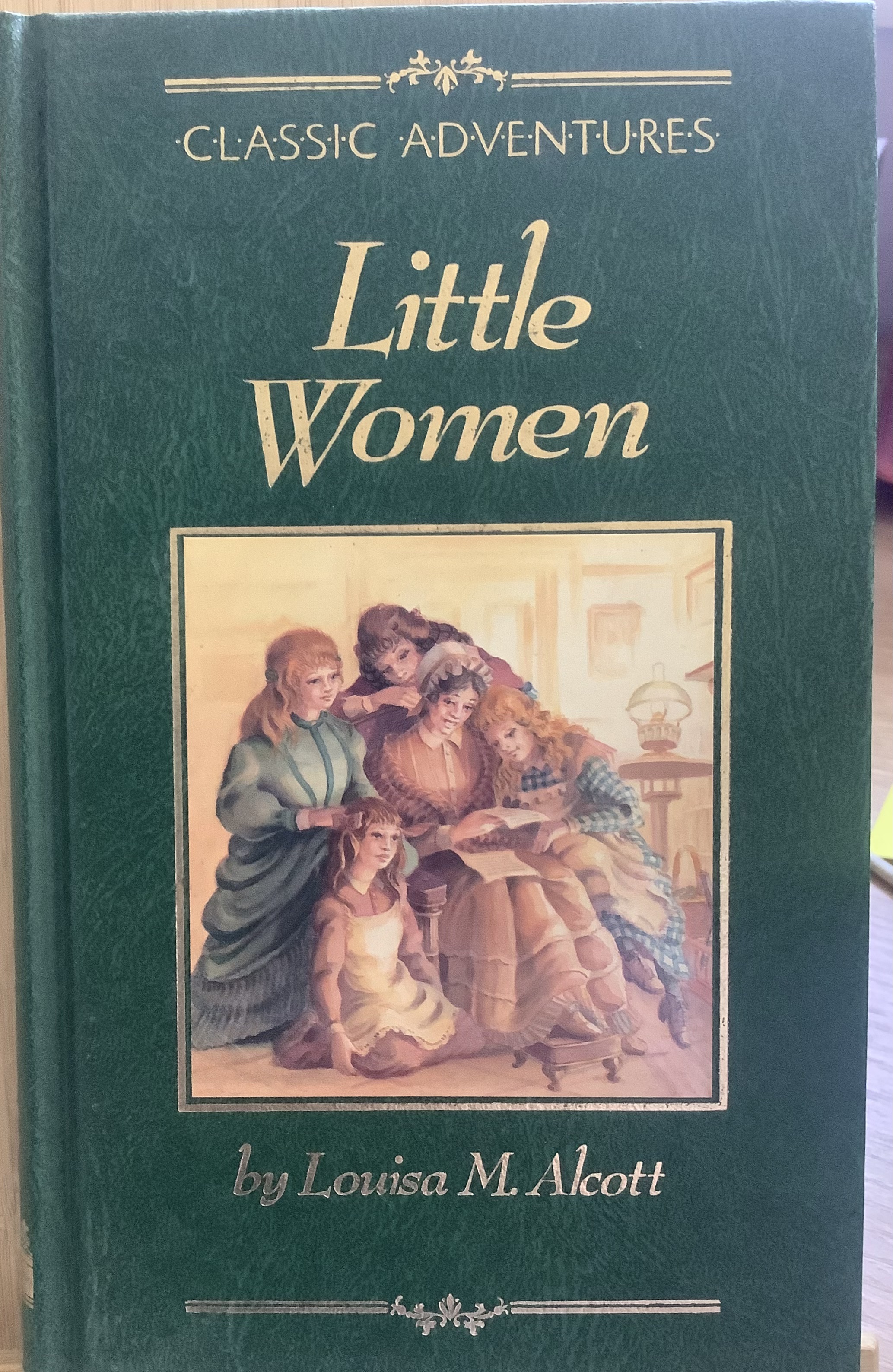 green hardcover novel. Little Women and author's name are in gold lettering. Illustration shows Marmee on a chair with all five girls surrounding her.